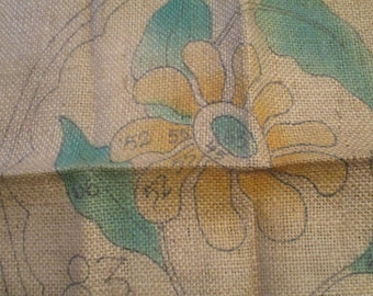 May include: Close-up of a burlap fabric with a floral design. The design features a large flower with yellow petals, a blue center, and teal leaves. The fabric has a rough texture and visible weave. Numbers are visible on the design.