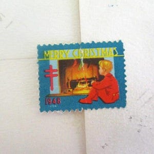 Op de afbeelding: Een vintage kerstpostzegel met een blauwe rand. De postzegel toont de woorden "MERRY CHRISTMAS" boven een afbeelding van een kind in rode kleding dat bij een open haard zit. Het jaartal "1946" staat onderaan gedrukt.