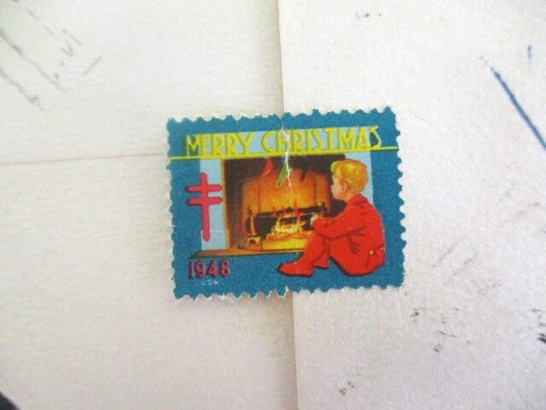 Op de afbeelding: Een vintage kerstpostzegel met een blauwe rand. De postzegel toont de woorden "MERRY CHRISTMAS" boven een afbeelding van een kind in rode kleding dat bij een open haard zit. Het jaartal "1946" staat onderaan gedrukt.