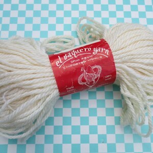 May include: Two skeins of off-white yarn with a red label that reads "el vaquero yarn" and "spun from imported wools". The yarn is 100% wool and has a triple twist. The background is a blue and white checkered pattern.