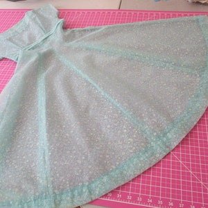 May include: A light blue, semi-sheer garment with a floral pattern, possibly a cape or dress. The fabric is laid out on a pink cutting mat. The garment has a collar and sleeves, with a rounded, flared shape.