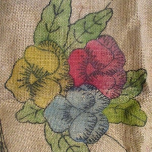 K&ouml;nnte beinhalten: Nahaufnahme eines Jutestoffs mit floralem Design. Das Design zeigt eine Ansammlung von Blumen in Gelb, Rot und Blau mit gr&uuml;nen Bl&auml;ttern. Die Blumen sind handgemalt mit schwarzen Umrissen, was eine rustikale, Vintage-&Auml;sthetik erzeugt.