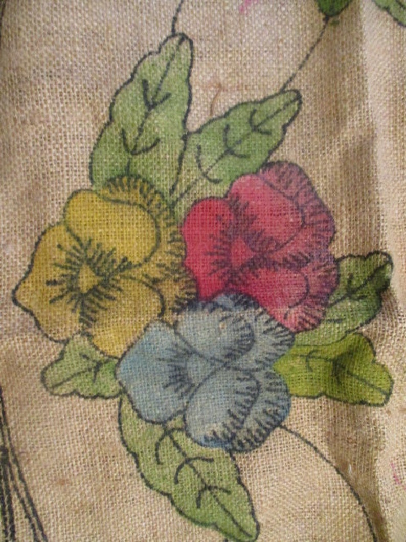 K&ouml;nnte beinhalten: Nahaufnahme eines Jutestoffs mit floralem Design. Das Design zeigt eine Ansammlung von Blumen in Gelb, Rot und Blau mit gr&uuml;nen Bl&auml;ttern. Die Blumen sind handgemalt mit schwarzen Umrissen, was eine rustikale, Vintage-&Auml;sthetik erzeugt.