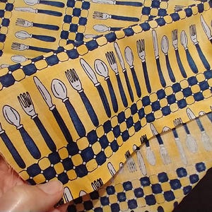 May include: Yellow fabric featuring a repeating pattern of white and navy blue cutlery, including forks, knives, and spoons. A navy and yellow chequered border frames the design. The fabric is folded, showing the pattern.