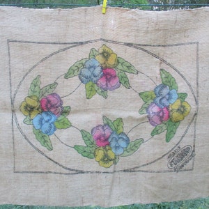 K&ouml;nnte beinhalten: Ein beiges Jutegewebe mit einem floralen Design. Es zeigt bunte Stiefm&uuml;tterchen in Gelb, Blau und Rosa, umgeben von gr&uuml;nen Bl&auml;ttern. Das Design ist von einem schwarzen Oval und einem rechteckigen Rahmen umschlossen. Der Text "STANDARD STAMPING CO." ist sichtbar.