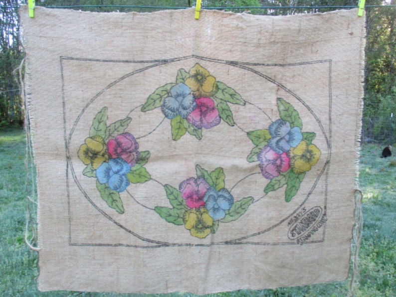 K&ouml;nnte beinhalten: Ein beiges Jutegewebe mit einem floralen Design. Es zeigt bunte Stiefm&uuml;tterchen in Gelb, Blau und Rosa, umgeben von gr&uuml;nen Bl&auml;ttern. Das Design ist von einem schwarzen Oval und einem rechteckigen Rahmen umschlossen. Der Text "STANDARD STAMPING CO." ist sichtbar.