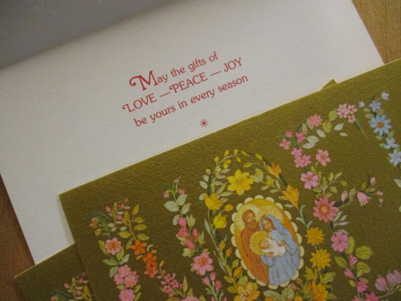 Puede incluir: Una tarjeta de Navidad con un dise&ntilde;o floral y el texto "Que los dones de AMOR - PAZ - ALEGR&Iacute;A sean tuyos en cada estaci&oacute;n." La tarjeta presenta una ilustraci&oacute;n de la Sagrada Familia.