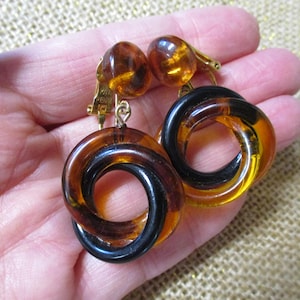 May include: A pair of clip-on earrings featuring a gold-tone metal mechanism with a round amber-colored bead at the top. Each earring has a dangling circular pendant composed of two interlocking rings in translucent dark brown and amber resin.