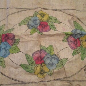 K&ouml;nnte beinhalten: Vintage Blumenstickmuster auf Jute. Das Design zeigt bunte Stiefm&uuml;tterchen in Gelb-, Blau- und Rott&ouml;nen, umgeben von gr&uuml;nen Bl&auml;ttern. Die Blumen sind in einem ovalen Rahmen mit schwarzer Umrandung angeordnet. Die Jute hat ein leicht gealtertes Aussehen.