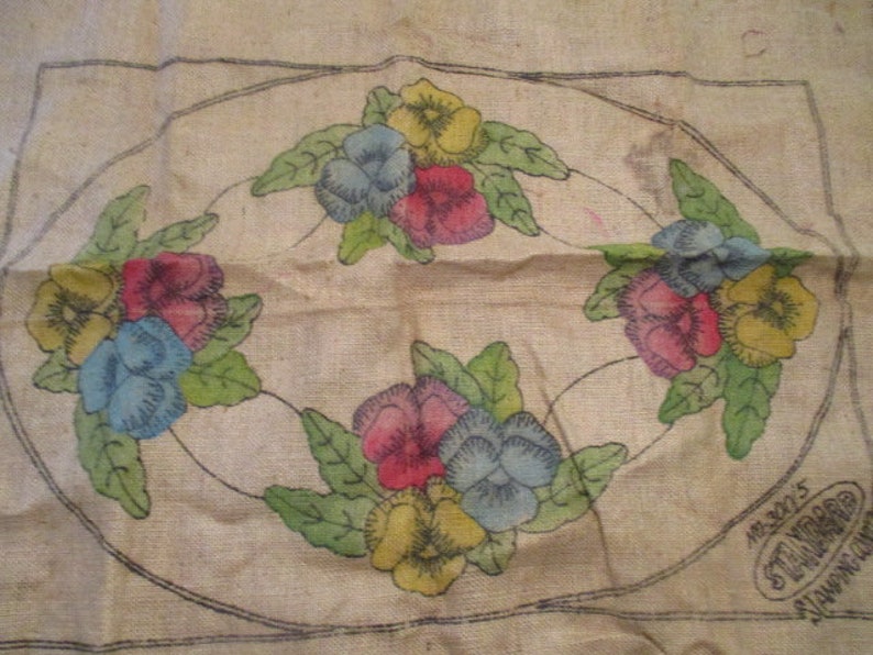 K&ouml;nnte beinhalten: Vintage Blumenstickmuster auf Jute. Das Design zeigt bunte Stiefm&uuml;tterchen in Gelb-, Blau- und Rott&ouml;nen, umgeben von gr&uuml;nen Bl&auml;ttern. Die Blumen sind in einem ovalen Rahmen mit schwarzer Umrandung angeordnet. Die Jute hat ein leicht gealtertes Aussehen.