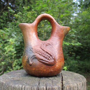 May include: A terracotta-colored ceramic vessel featuring a double-spout design connected by a central arched handle. The surface displays a textured, embossed relief of a corn cob on the front, with dark, mottled shading across the clay body.
