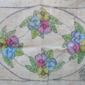 K&ouml;nnte beinhalten: Vintage Blumenstickmuster auf beigem Stoff. Das Design zeigt vier Gruppen von Stiefm&uuml;tterchen in Gelb, Rosa und Blau, umgeben von gr&uuml;nen Bl&auml;ttern, in einem ovalen Rahmen. Der Text "STANDARD STAMPING CO." ist sichtbar.
