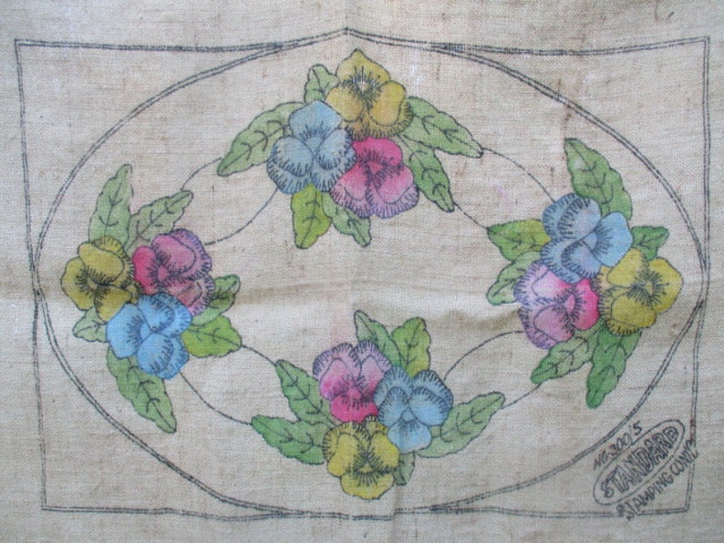 K&ouml;nnte beinhalten: Vintage Blumenstickmuster auf beigem Stoff. Das Design zeigt vier Gruppen von Stiefm&uuml;tterchen in Gelb, Rosa und Blau, umgeben von gr&uuml;nen Bl&auml;ttern, in einem ovalen Rahmen. Der Text "STANDARD STAMPING CO." ist sichtbar.