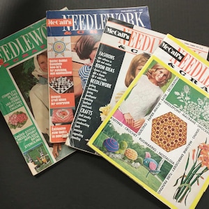 May include: A stack of vintage "Needlework & Crafts" magazines. The covers feature images of women, craft projects, and text. The magazines are in various colors, including green, yellow, and black, and are stacked on a dark surface.