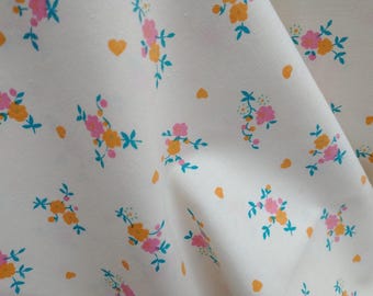 May include: Off-white fabric featuring a repeating pattern of small floral bouquets in pink and orange, with teal leaves. Orange heart shapes are scattered across the design. The fabric is creased, indicating it is a textile.