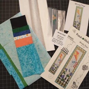 Könnte beinhalten: Ein Patchwork-Set mit Stoffstücken in Blau, Grün, Orange und Schwarz. Das Set enthält ein Muster mit dem Titel "Windy Days" von Patch Abilities, Inc. Die fertige Größe beträgt 15 cm x 56 cm.