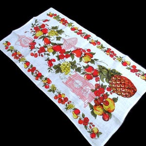 May include: White rectangular kitchen towel with a colorful fruit design. The towel features illustrations of red apples, yellow peaches, green grapes, and a pineapple. Pink decorative elements are also present.