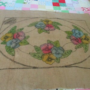 K&ouml;nnte beinhalten: Beige Jute-Textil mit floralem Design. Das Design zeigt eine ovale Anordnung bunter Stiefm&uuml;tterchen in Gelb, Rosa und Blau, umgeben von gr&uuml;nen Bl&auml;ttern. Die Umrisse sind schwarz.