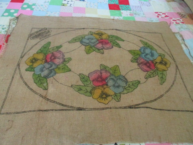 K&ouml;nnte beinhalten: Beige Jute-Textil mit floralem Design. Das Design zeigt eine ovale Anordnung bunter Stiefm&uuml;tterchen in Gelb, Rosa und Blau, umgeben von gr&uuml;nen Bl&auml;ttern. Die Umrisse sind schwarz.