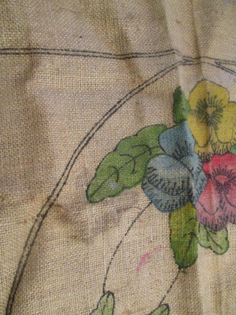 K&ouml;nnte beinhalten: Nahaufnahme eines Jutestoffs mit handgemaltem Blumendesign. Die Blumen sind in Gelb-, Blau- und Rosat&ouml;nen gehalten, mit gr&uuml;nen Bl&auml;ttern. Schwarze Umrisse definieren die Blumenformen und -stiele. Der Stoff hat ein raues, strukturiertes Aussehen.