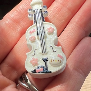 May include: A white violin-shaped charm with pink flower accents and a Hello Kitty face. The miniature instrument has black strings and purple details, held in a hand.