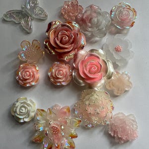 May include: A collection of iridescent floral and butterfly embellishments in various sizes and shades of pink, white, and peach. The decorative elements include roses, a multi-petal flower, and a butterfly.