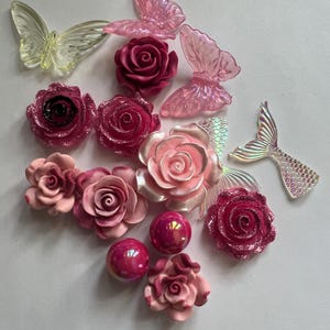 May include: Assortment of decorative embellishments including pink roses, iridescent mermaid tails, and translucent butterflies. The roses vary in shades of pink and red, some with glitter accents. The butterflies are light yellow and pink.