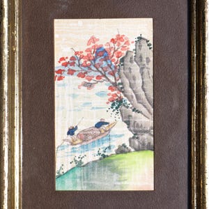 May include: Framed watercolor painting depicting a serene river scene. A boat with two figures floats on the water, beneath a tree with red blossoms and a rocky cliff. The frame is gold-toned.