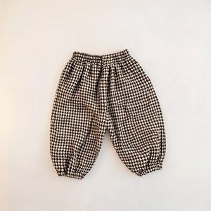 Puede incluir: Un pantalón con estampado de pata de gallo en blanco y negro. La prenda cuenta con una cintura elástica y puños elásticos en la parte inferior de cada pierna, creando una silueta fruncida y redondeada a lo largo de las piernas.