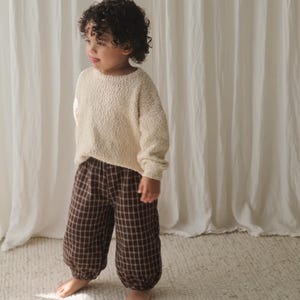 Puede incluir: Un suéter de manga larga en color crema con textura, combinado con pantalones marrones con un patrón de cuadrícula blanco. Los pantalones cuentan con una cintura elástica y puños fruncidos en los tobillos, creando una silueta holgada.