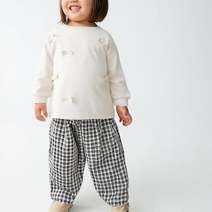 May include: A child's outfit featuring a cream-colored long-sleeved shirt adorned with small bow accents and black and white checkered pants. The outfit is paired with beige sneakers, creating a playful and comfortable look.