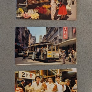 May include: Three vintage postcards featuring scenes from San Francisco. The first postcard shows a flower stand with a variety of flowers, a man and woman shopping, and a city street in the background. The second postcard shows a cable car traveling down a street in the city. The third postcard shows a fish market with three men holding crabs and a sign that says "2 for $1.00".
