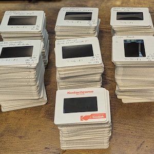 May include: Stacks of vintage Kodachrome slides with handwritten labels, featuring dates and locations like "Lake Granty" and "Ute Peak." The slides are arranged on a wooden surface, showcasing a collection of photographic memories.