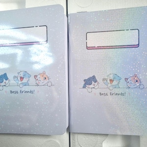 May include: Two notebooks with a shimmering, light blue cover. Each notebook features a rectangular label area and a row of three cartoon cats in blue, light blue, and orange. The text "Best friends!" is below the cats.