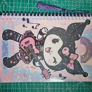 May include: Spiral-bound notebook with a cartoon character in black and white, holding a pink guitar. The notebook has a purple tassel and a cover with a colourful design, musical notes and the text "NOTEBOOK".