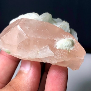 Może przedstawiać: Zbliżenie różowego kryształu kwarcu z białymi i zielonymi osadami mineralnymi. Kwarc ma fasetowaną, krystaliczną strukturę. Ciemne tło podkreśla naturalne kolory i tekstury okazu mineralnego.