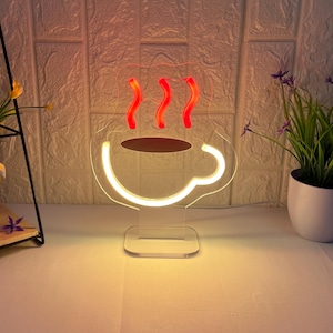 May include: A neon sign in the shape of a coffee cup. The cup outline and steam are illuminated in white and red, respectively. The coffee inside the cup is brown. The sign is on a clear base.
