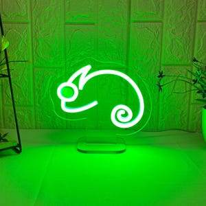 May include: A neon green chameleon-shaped light fixture. The light is a bright, glowing green, with a clear base. The background is a light green brick wall. There are two green potted plants on either side of the light.
