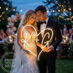May include: A neon sign in the shape of a heart with the letters "R + G" in a cursive font. The sign is held by a couple at an outdoor wedding. The sign is lit with a warm, bright light. The words "BRIGHT YOUR NEON" are in the lower left corner.