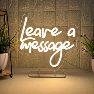 Puede incluir: Un letrero de neón blanco que dice "Leave a message" en una fuente cursiva. El letrero está montado sobre una base de acrílico transparente. El fondo es una pared texturizada. Hay plantas en macetas a ambos lados.