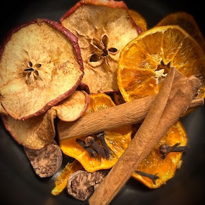 May include: A collection of dried food items including sliced red apples, bright orange citrus slices, whole cinnamon sticks, and dark brown cloves. The ingredients are arranged together, showcasing varied textures and warm, earthy colors.