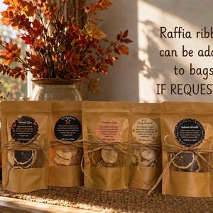 May include: Five brown kraft paper pouches with clear windows containing dried fruit slices, each tied with twine and a circular label. Text on the right reads: Raffia ribbon can be added to bags IF REQUESTED. The pouches feature labels for Harvest Glow, Fall Bonfires, Pumpkin Spice, Candy Cornucopia, and Autumn Wreath.