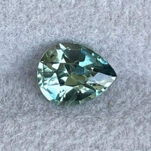 May include: A pear-shaped, faceted gemstone with a light teal color. The gem has a brilliant cut, reflecting light and creating a sparkling effect. The stone is set against a light gray, textured background.