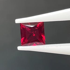 Könnte beinhalten: Ein quadratisch geschliffener, tiefroter Edelstein wird von einer weißen Pinzette vor einem dunklen Hintergrund gehalten. Die Facetten des Edelsteins reflektieren das Licht und zeigen seine lebendige Farbe und Klarheit. Es ist wahrscheinlich ein Rubin.