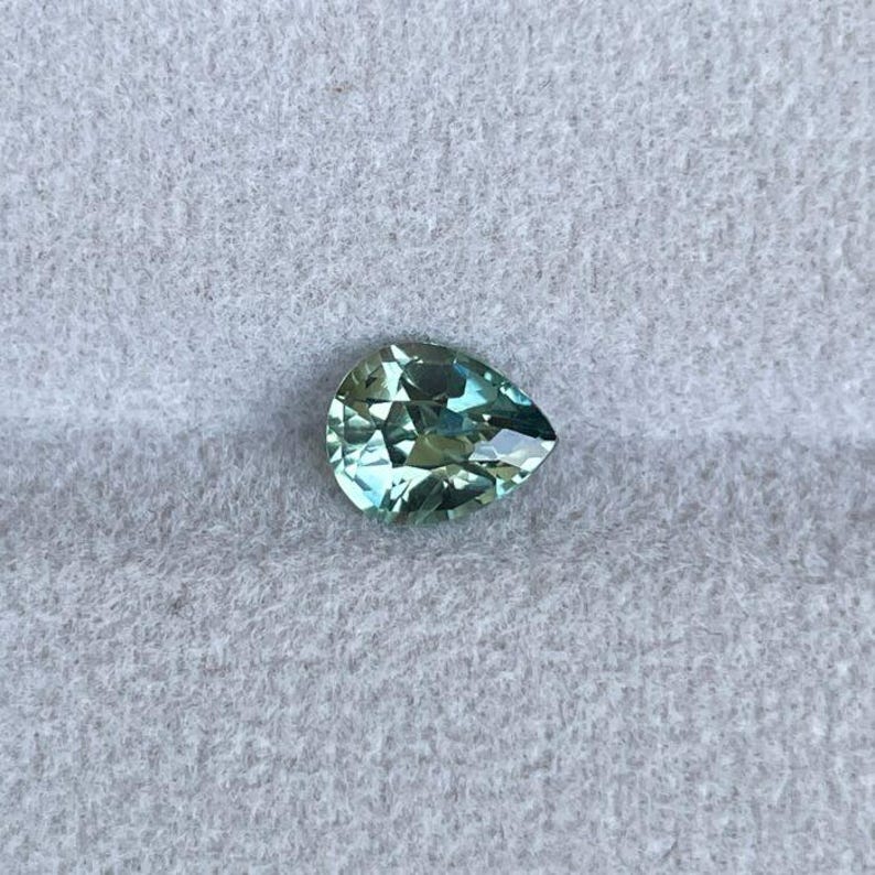 May include: A pear-shaped, faceted gemstone with a light teal color. The gem has a brilliant cut, reflecting light and creating a sparkling effect. The stone is set against a light gray, textured background.