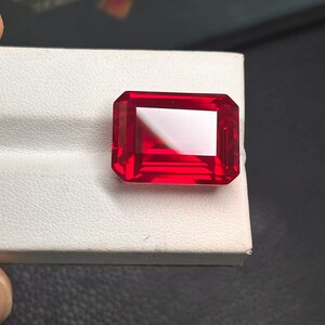 May include: A vibrant, rectangular-cut, red gemstone. The gem is set against a white surface, showcasing its clarity and rich color. The cut features precise facets, reflecting light. The gemstone is a striking example of fine jewelry.