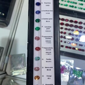 May include: A display case showcasing a variety of gemstones, including red ruby, royal blue, cornflower blue, Zambian green, and others. Each gem is labeled with its name and color, offering a spectrum of hues from pink to yellow.