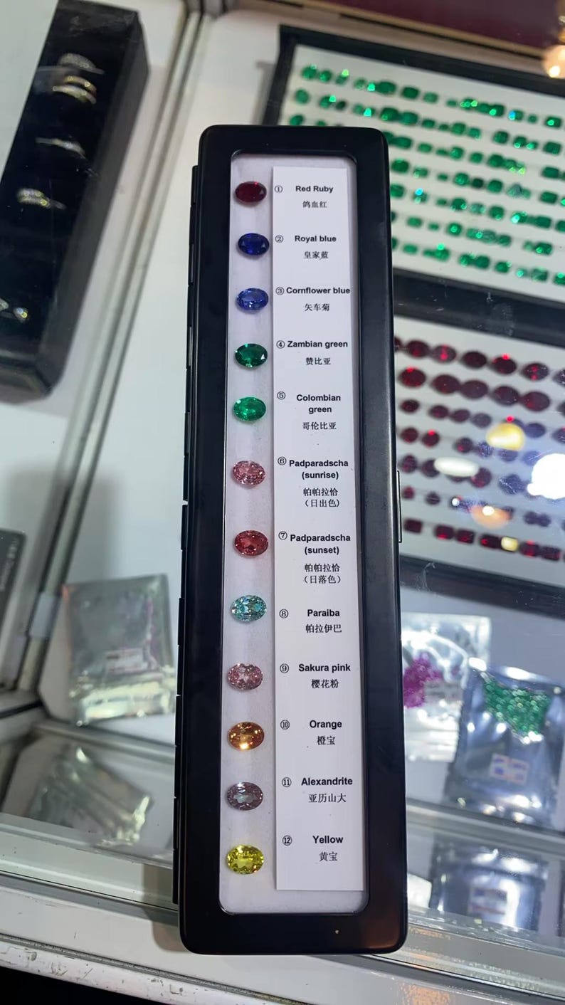 May include: A display case showcasing a variety of gemstones, including red ruby, royal blue, cornflower blue, Zambian green, and others. Each gem is labeled with its name and color, offering a spectrum of hues from pink to yellow.