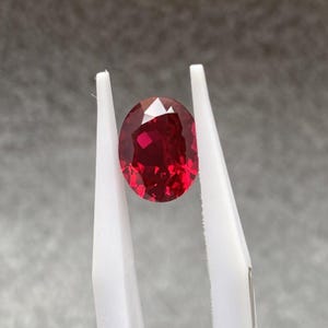 Pode incluir: Uma gema facetada, de forma oval, vermelho escuro, segurada por pinças brancas. A gema tem um corte brilhante, refletindo a luz. O fundo é cinza suave, destacando a cor vibrante da rubi. Esta imagem mostra uma pedra preciosa.