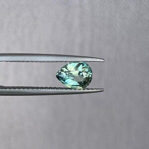 May include: A pear-shaped, faceted gemstone with shades of teal and green, held by a pair of silver tweezers. The gem has a brilliant cut, reflecting light. The background is a plain, light gray.
